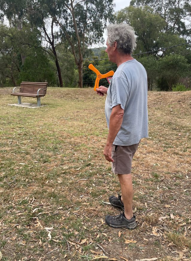 Man holding an orange boomerang in a park with trees and a bench in the background